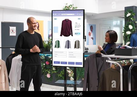 Male customer is shown clothing on an interactive kiosk board by female assistant in mall clothing store. Asian retail employee assisting a client who is self-ordering modern fashion items. Stock Photo