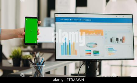 Woman holding phone with greenscreen at business office desk, startup employee looking at blank copyspace displa on smartphone. Person working with isolated design template on phone. Stock Photo