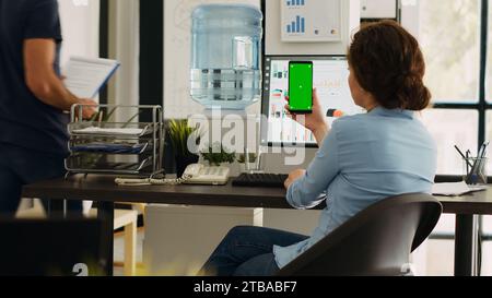 Businesswoman looks at greenscreen on phone while she checks marketing renevue in coworking space, employee holding mobile phone display showing isolated copyspace template. Specialist at job. Stock Photo