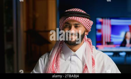 Portrait of smiling Arab freelancer working from home, taking a break after typing on his laptop all day, close up. Middle eastern man in traditional attire wearing headscarf, being productive Stock Photo