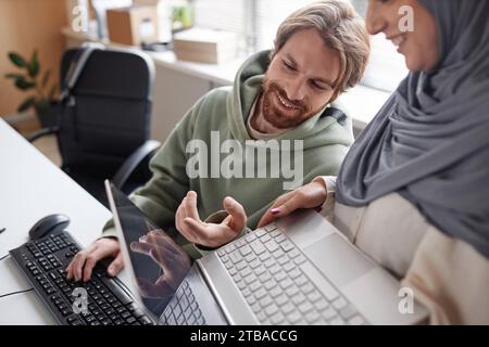 Portrait of bearded young man talking to muslim colleague in IT office and smiling while pointing at laptop screen Stock Photo