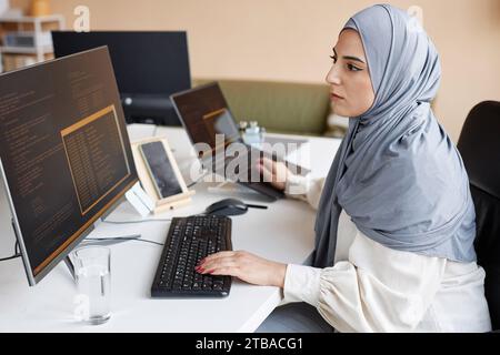 Side view portrait of Muslim young woman wearing modest clothing in office while working as female software developer and writing code with multiple computer devices Stock Photo