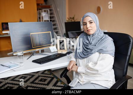 Portrait of Muslim young woman as female IT programmer sitting at workplace with computers and looking at camera Stock Photo