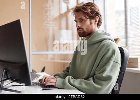 Side view portrait of bearded young man wearing hoodie and using computer in IT development office, copy space Stock Photo