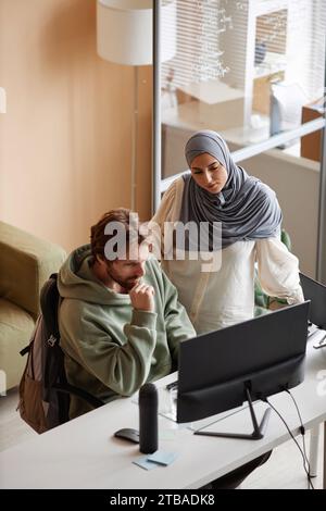 Vertical high angle portrait of two young people discussing project in office with Muslim young woman looking at computer screen Stock Photo