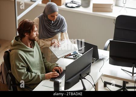 High angle portrait of female computer programmer wearing headscarf in office and showing laptop screen to colleague during code review Stock Photo