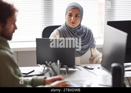 Front view portrait of young Middle Eastern woman looking at laptop screen while working with multiple computer devices in office Stock Photo