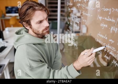 Side view at bearded young man writing data code ideas on glass in office while developing new technology Stock Photo