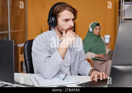 Side view portrait of bearded IT professional wearing headphones at workplace and looking at computer screen with focused face expression Stock Photo