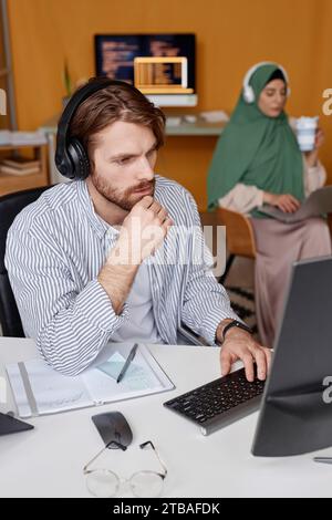Vertical portrait of bearded young man wearing headphones and looking at computer screen while writing code in IT office Stock Photo