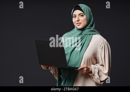 Waist up portrait of confident Muslim woman holding open laptop and smiling at camera as female IT professional, copy space Stock Photo