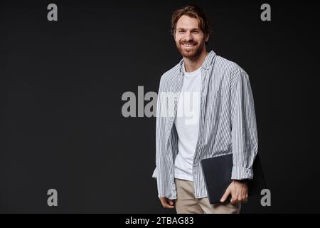Portrait of smiling bearded man holding laptop and smiling at camera while standing on black background in studio, copy space Stock Photo