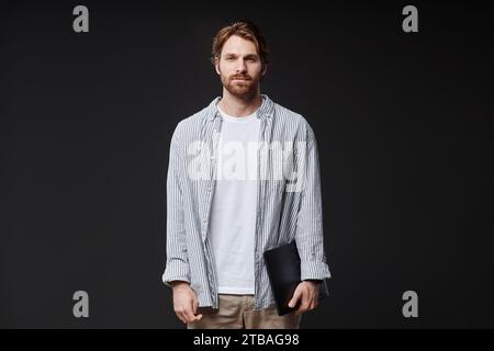 Front view portrait of bearded young man holding laptop and looking at camera standing on black background in studio, copy space Stock Photo
