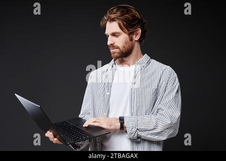 Waist up portrait of young bearded man holding open laptop while standing on black background in studio, copy space Stock Photo