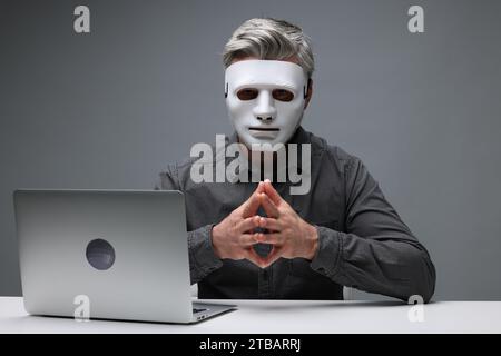 Man in mask sitting at white table with laptop against grey background Stock Photo