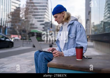 Fashionable guy sitting in urban street searching info in laptop computer, working, posting photos Stock Photo