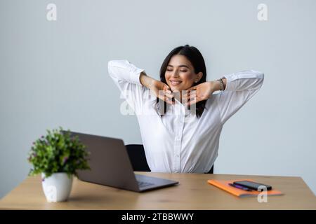Happy female office worker leaning back on chair, relaxing after successful work at her workplace in office, resting with hands behind head Stock Photo