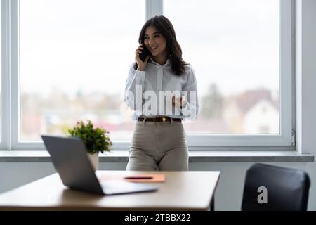Young brunette woman talking on mobile phone while standing in modern office Stock Photo