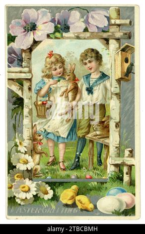 Original, charming Edwardian era Easter greetings card, brother and sister, girl is dressed like Alice in Wonderland, the boy wears a typical sailor suit popular at this time. There are eggs in a basket, chicks, hares or rabbits. floral border. Sent from Brooklyn, U.S.A. Circa 1905 / 1910. Stock Photo