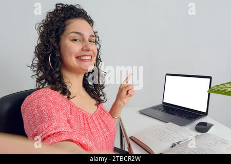 Selfie portrait of young Latin Venezuelan woman sitting in her office smiling and pointing at laptop screen with her finger, copy space. Stock Photo