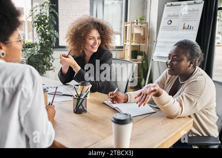 happy multicultural businesswomen discussing startup near notebooks and coffee to go, creative team Stock Photo