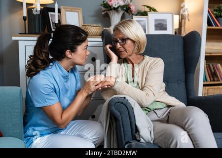 Young woman doctor health care worker medic nurse comforting older senior woman at home visit, patient suffering from clinical depression and anxiety. Stock Photo