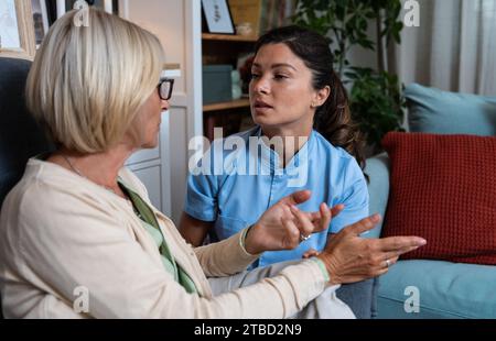 Young woman doctor health care worker medic nurse comforting older senior woman at home visit, patient suffering from clinical depression and anxiety. Stock Photo
