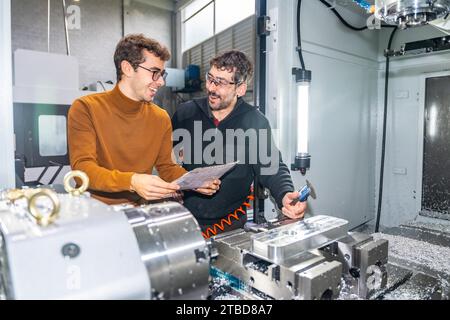 Two engineers talking distracted while examining a piece of metal next to a milling machine in a cnc factory Stock Photo