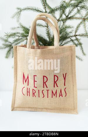 This is a photo of a burlap tote bag with Merry Christmas written in red on it, sitting in front of a small Christmas tree. Stock Photo