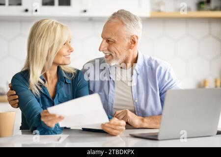 Portrait Of Cheerful Elderly Spouses Sitting In Kitchen And Checking Financial Papers Stock Photo