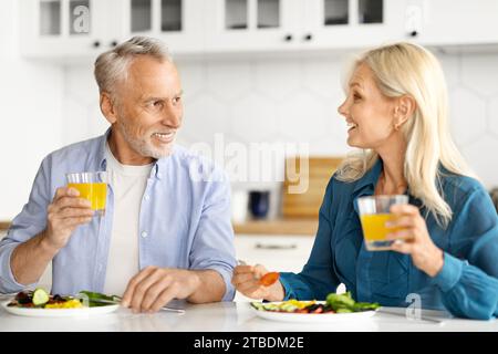 Happy Senior Spouses Chatting And Drinking Orange Juice During Breakfast In Kitchen Stock Photo