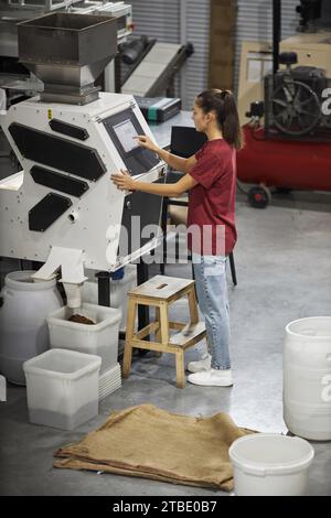 Vertical full length portrait of young woman operating machines at food factory and using touch screen control panel Stock Photo