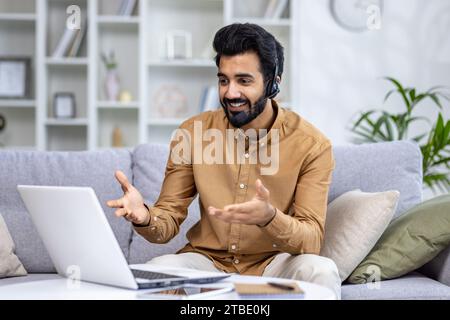Hindu businessman in a headset sits on a sofa in front of a laptop, has a work video call, consults a client, a student has an online class. Stock Photo
