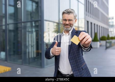 Happy senior businessman holding out card and showing thumbs up like, standing near office, smiling and looking at camera. Stock Photo