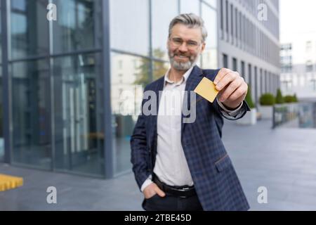 A smiling businessman in a suit holds a card in his hands, stands on the street in front of the office, looks at the camera. Stock Photo