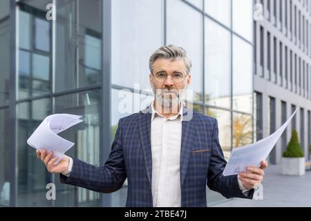 Confused senior businessman holds documents in hands, spreads hands, problems in business, looks at camera. Stock Photo