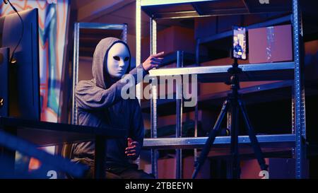 Hacker wearing anonymous mask doing ransom video after gathering information from users computers using spyware software. Cybercriminal blackmailing victim in graffiti painted hideout Stock Photo