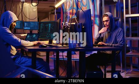 Panning shot of hackers working hard on writing lines of code on hideout computers, trying all methods of getting past security systems. Professional scammers building scripts that can hack devices Stock Photo