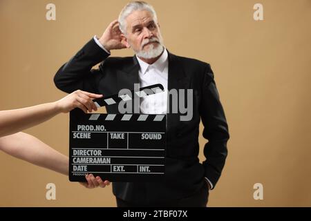 Senior actor performing role while second assistant camera holding clapperboard on beige background, selective focus. Film industry Stock Photo