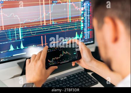 Stock exchange, trading.Male trader sits in home office in front of screens with graphs and financial charts, using smartphone, predicts, analyzing and studying the growth or decline of cryptocurrency Stock Photo