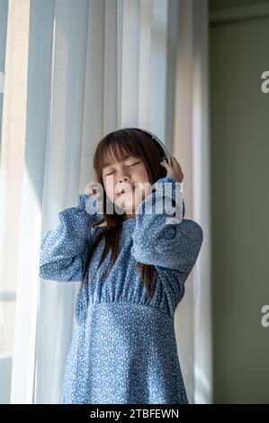 A cute and happy young Asian girl in a blue dress is standing by the window and enjoying listening to music on headphones in the living room. Kid and Stock Photo