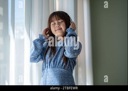 A cute and happy young Asian girl in a blue dress is standing by the window and enjoying listening to music on headphones in the living room. Kid and Stock Photo