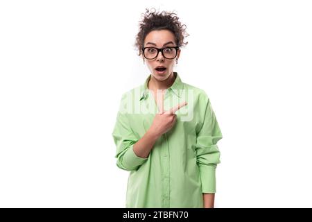young surprised brunette businesswoman with curly hair dressed in a green shirt points her hand to the side Stock Photo