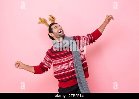 Christmas studio shot portrait of cheerful Caucasian man wearing sweater and fancy reindeer headband laughing and dancing in pink color isolated backg Stock Photo