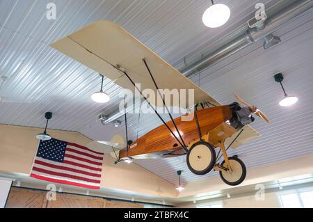 A small tan model biplane with an orange wooden fuselage and spoked wheels hangs from the ceiling above an American flag in a museum hall in Ohio. Stock Photo