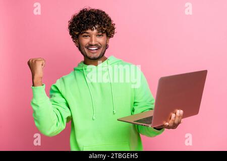 Photo of successful startup investor trader indian young guy fist up working freelance using netbook isolated on pink color background Stock Photo