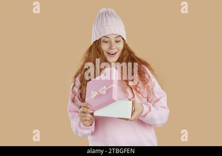 Amazed redhead girl with curly hair opening a present gift box and smiling on beige background. Stock Photo
