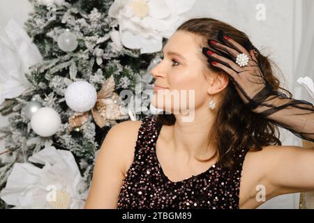 Smiling woman in glitter blouse and gloves in profile near Christmas tree Stock Photo