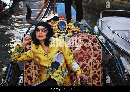 A confident woman wearing a golden oriental Crane coat in a gondola being rowed by venetian gondolier through picturesque canals of Venice, Italy Stock Photo