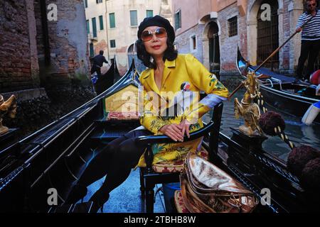 Venice, Italy, an elegant mature woman wearing a golden oriental coat sitting in a gondola being rowed past palazzo, and other gondolier & gondolas Stock Photo